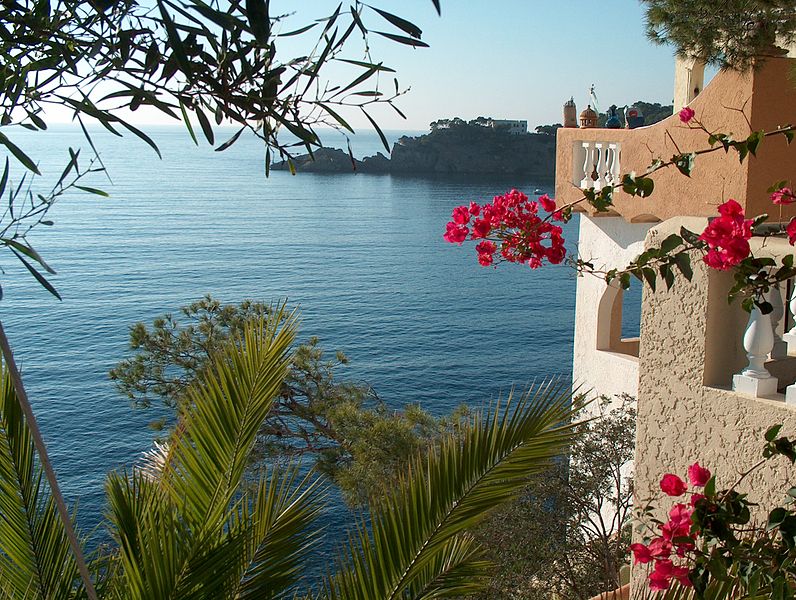 A vista costeira com flores rosas brilhantes, um mar azul, um edifício de areia e uma costa rochosa distante sob um céu limpo oferecem o cenário perfeito enquanto você explora como chegar a Valência.