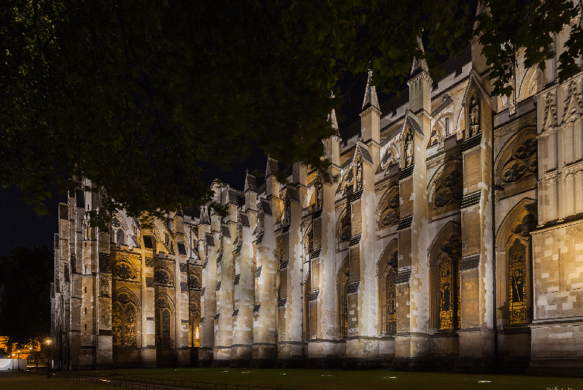 Fachada da catedral em estilo gótico iluminada à noite, cercada por árvores, oferece uma das melhores atrações de Londres para os visitantes admirarem.