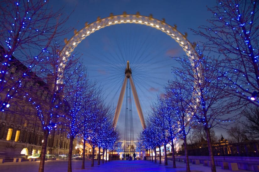 A London Eye, uma das melhores atrações de Londres, fica iluminada à noite, cercada por árvores adornadas com luzes azuis.