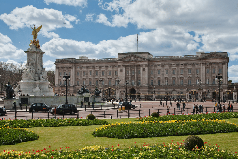 O Palácio de Buckingham, uma das melhores atrações de Londres, ergue-se majestosamente com uma estátua em primeiro plano. As pessoas passeiam tranquilamente pelos jardins coloridos sob um céu parcialmente nublado, capturando a essência da elegância e do charme britânicos.