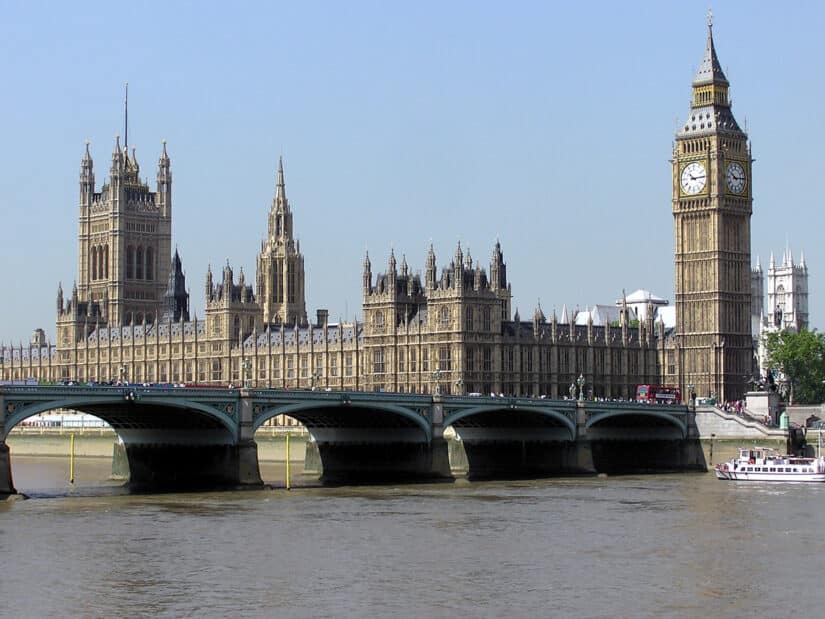 As Casas do Parlamento e o Big Ben, duas das melhores atrações de Londres, erguem-se majestosamente ao lado do Rio Tâmisa, sob um céu azul claro.