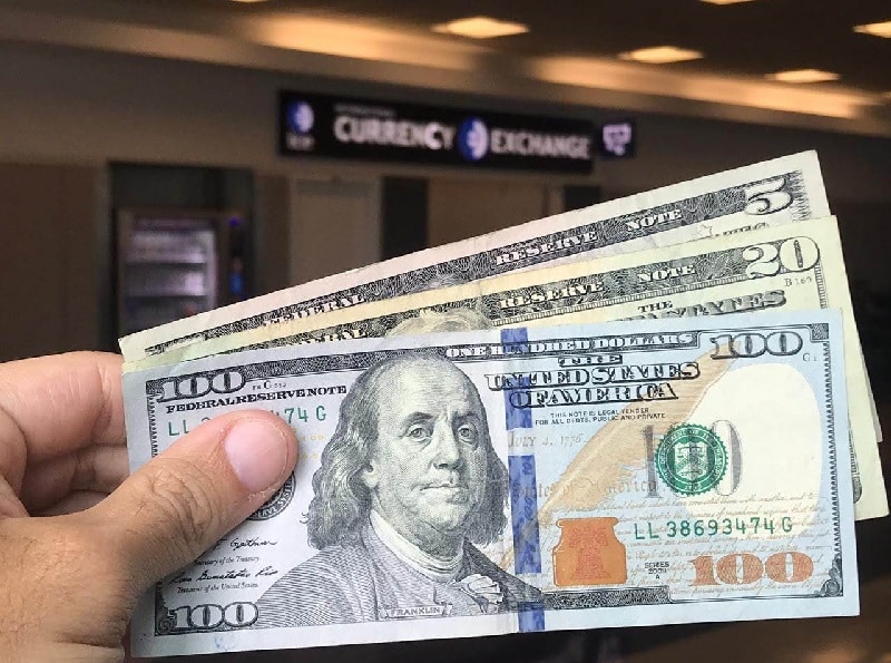 Uma mão segura firmemente uma moeda americana — notas de US$ 100, US$ 20 e US$ 5 — tendo como pano de fundo uma placa de câmbio, pronta para abastecer aventuras com dicas privilegiadas de Cancún.