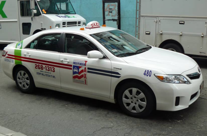 Um carro-táxi branco, com os logotipos da empresa e o número de contato 268-1313, está estacionado em uma movimentada rua da cidade, pronto para levar passageiros a destinos como o aeroporto de Boston.