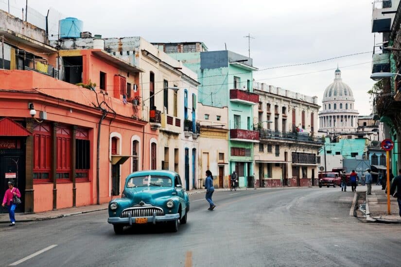 Um carro azul antigo passa por edifícios coloridos, evocando o charme de Cuba — não se esqueça de seu seguro viagem para Cuba.