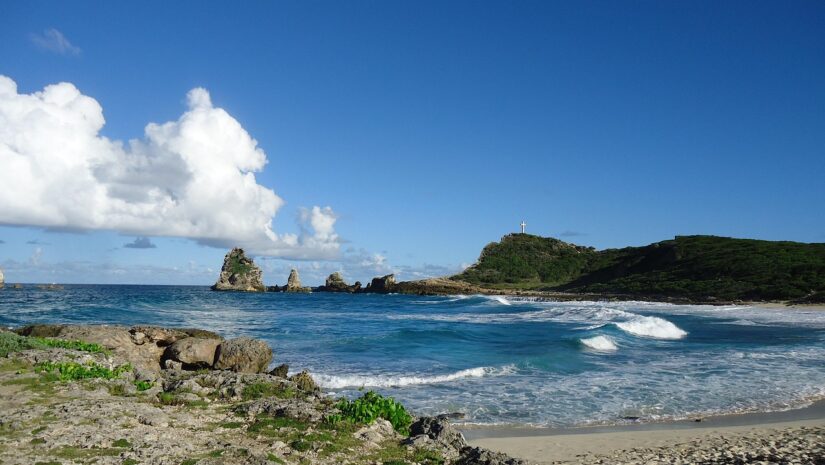 Praia rochosa com ondas azul-turquesa, morro verde e céu azul – ideal para uma viagem segura para Caribe e América Central.