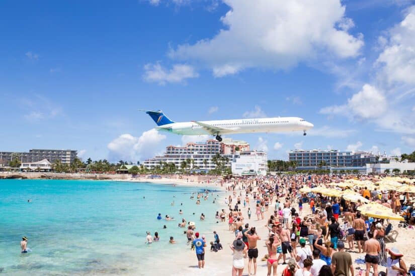 Uma grande multidão na praia observa um avião voando baixo perto da pista — considere seguro viagem para St Marteen/St Martin.