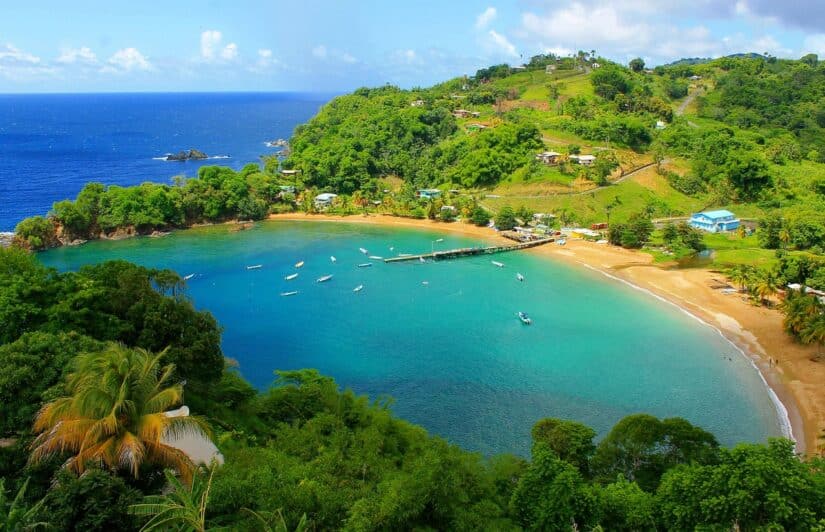 Uma baía azul-turquesa com barcos, praia de areia, colinas verdes e um lembrete de viagem segura para Trinidad e Tobago.