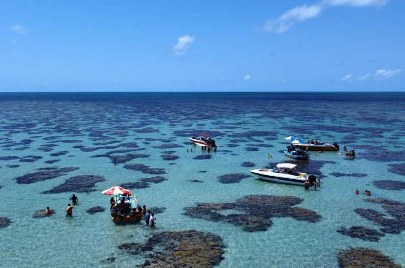 Pequenos barcos e pessoas desfrutam de passeios em Natal em águas claras e rasas com manchas de coral visíveis sob um céu azul.