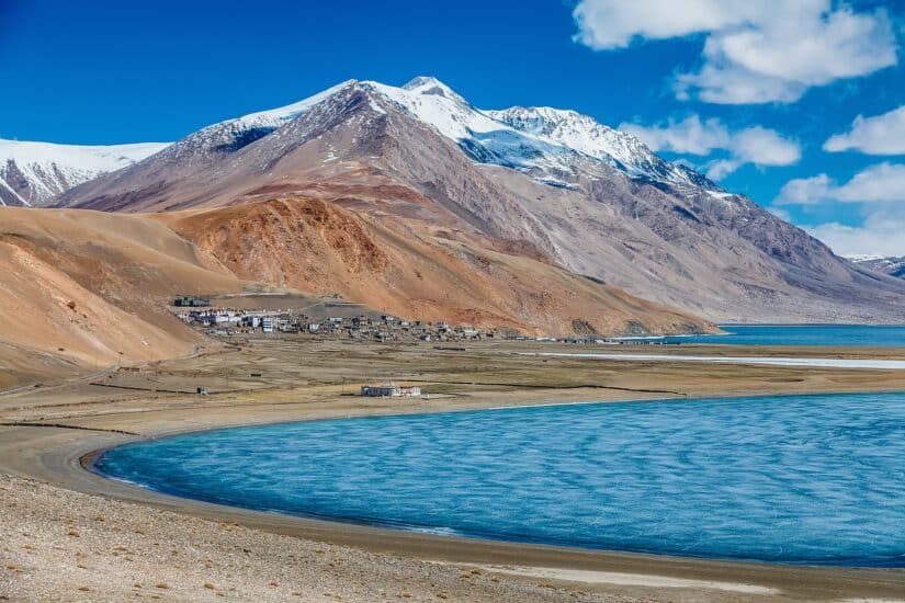 Paisagem montanhosa com picos nevados perto de uma pequena vila boliviana e um lago azul calmo sob um céu parcialmente nublado.