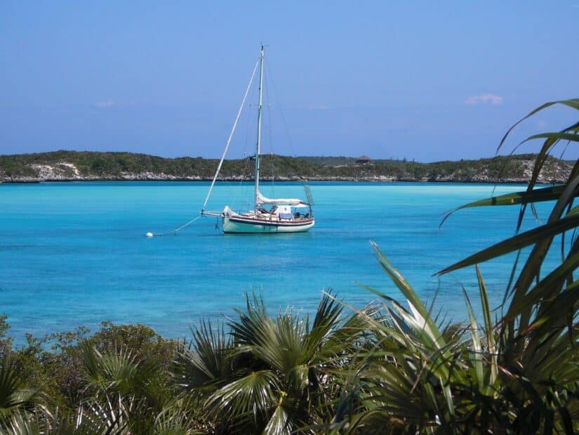 Veleiro ancorado nas águas azul-turquesa das Bahamas, com vegetação exuberante e uma costa distante sob um céu azul claro.