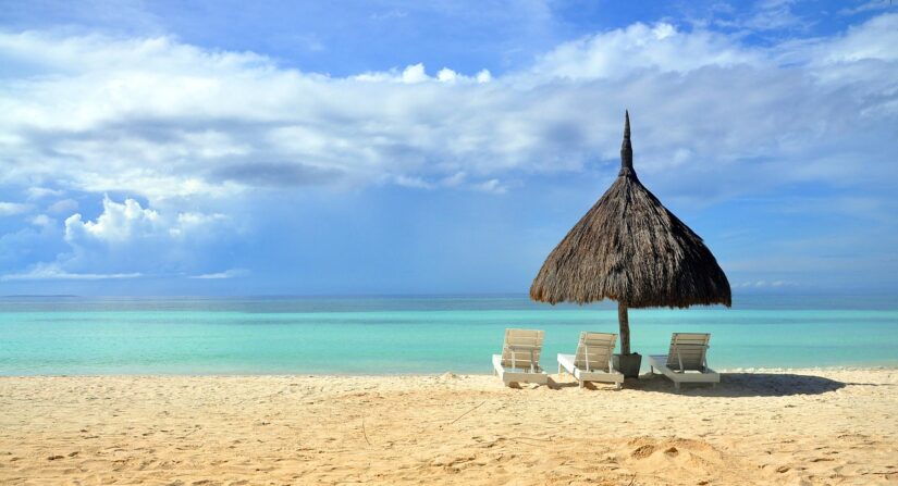 Três espreguiçadeiras sob um guarda-sol de palha em uma praia de areia com eSIM Filipinas, com vista para um oceano azul-turquesa e céu nublado.