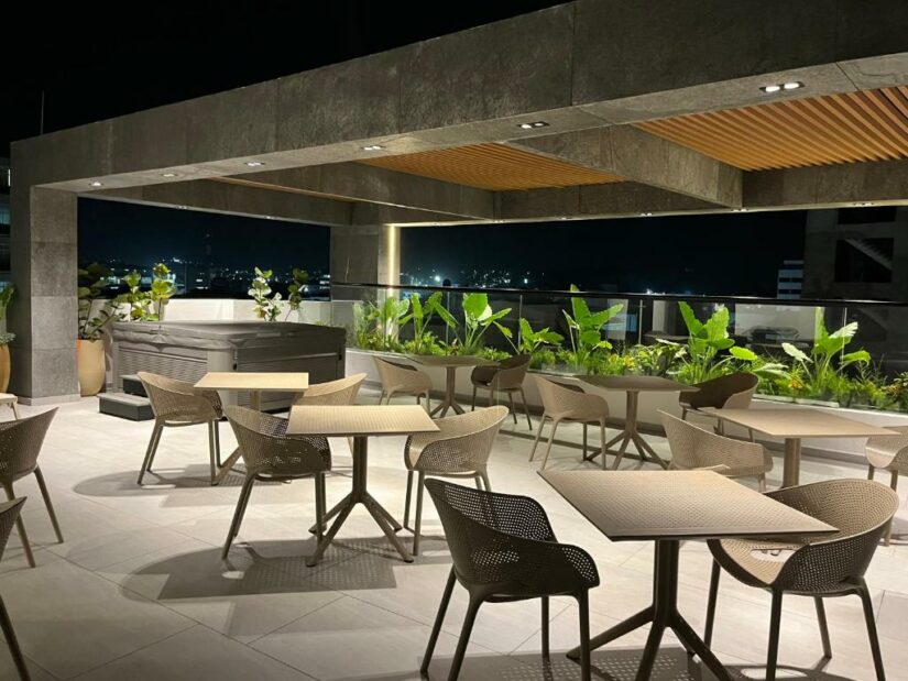 Terraço à noite em um hotel boutique, com mesas vazias, cadeiras, vegetação exuberante e luzes da cidade de San Andrés.