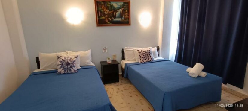 Duas camas com roupa de cama azul, uma pintura na parede e toalhas dobradas. Iluminação suave e cortinas escuras neste retiro em Cancún.
