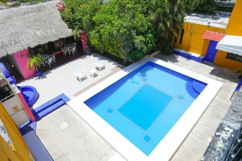 Vista aérea de uma piscina, pátio, paredes vibrantes e árvores. Perto, há um gazebo com telhado de palha em hotéis baratos em Cancún.