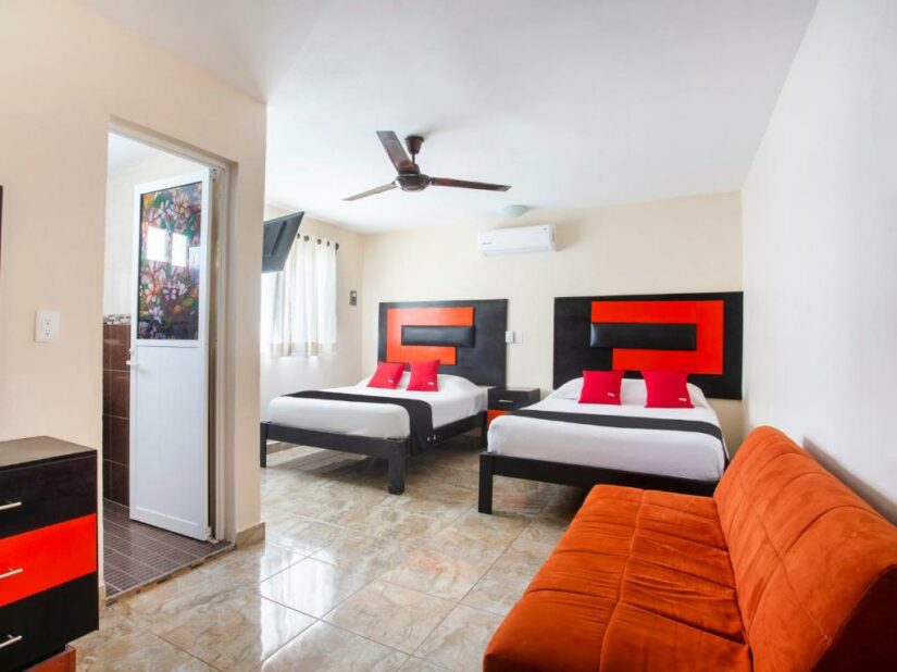 Quarto de hotel bem iluminado com duas camas, detalhes em vermelho, sofá laranja, ventilador de teto e uma porta no banheiro — uma joia entre os hotéis baratos em Cancún.