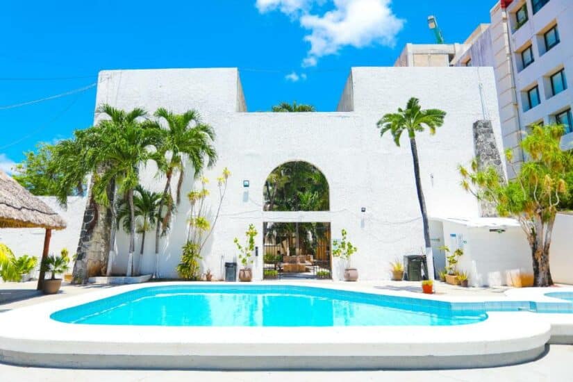 Uma área de piscina ensolarada em um edifício branco com palmeiras e um arco, oferecendo hotéis baratos em Cancún com preços acessíveis e tranquilidade.