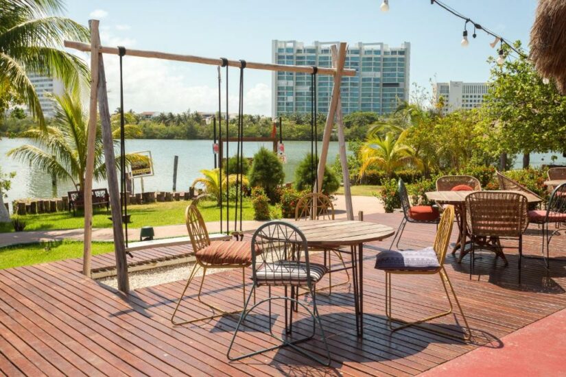 Pátio ao ar livre com deck de madeira, balanços e assentos, com vista para um lago com edifícios; perfeito para aproveitar hotéis baratos em Cancún.
