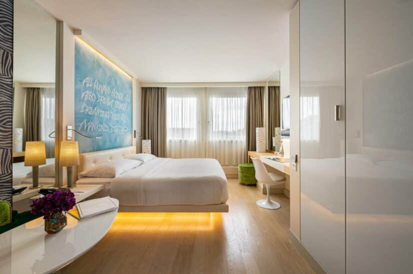 Quarto de hotel moderno em Genebra com cama, arte na parede, escrivaninha, cadeira e iluminação aconchegante. Decoração branca e luz natural vinda das janelas.