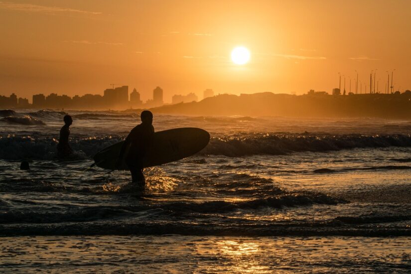 Os surfistas entram no oceano ao pôr do sol, com o horizonte da cidade e hotéis em Punta del Este de frente para o mar ao fundo.