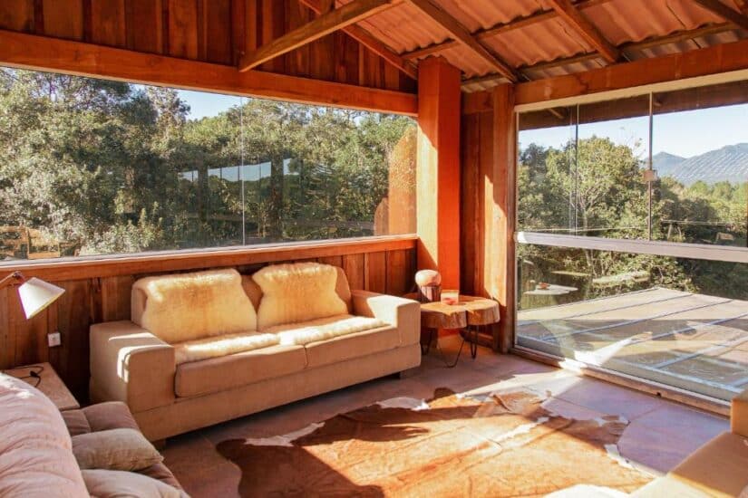 Interior de um chalé com três sofás, luminária ao lado, tapete e uma varanda com vista para a vegetação