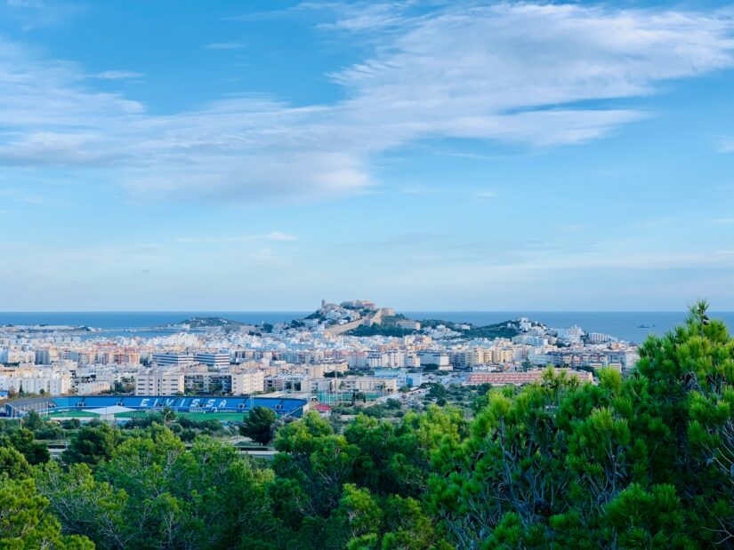 melhores hotéis em Dalt Vila nas Ilhas Baleares Paisagem urbana com um castelo no topo de uma colina, perfeita para explorar onde ficar em Ibiza, com árvores e mar ao fundo.