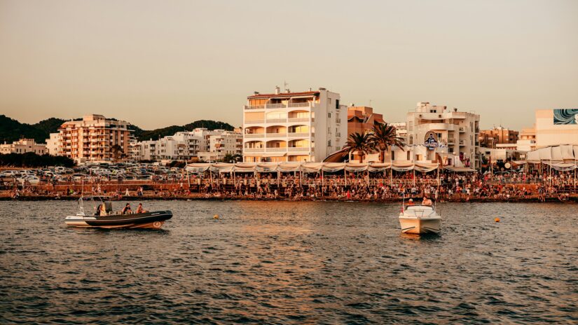 onde ficar em Ibiza na Espanha Dois barcos na água ao pôr do sol, perto das Ilhas Baleares, com multidões e prédios brancos ao longo da costa.