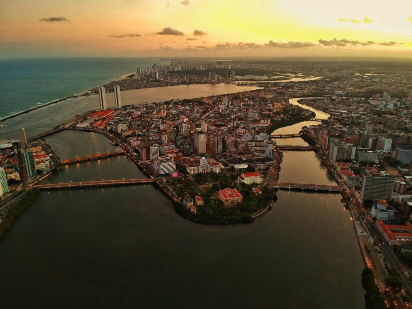 Vista aérea de Recife ao pôr do sol, mostrando onde ficar em Recife com rios, prédios altos e litoral cênico.