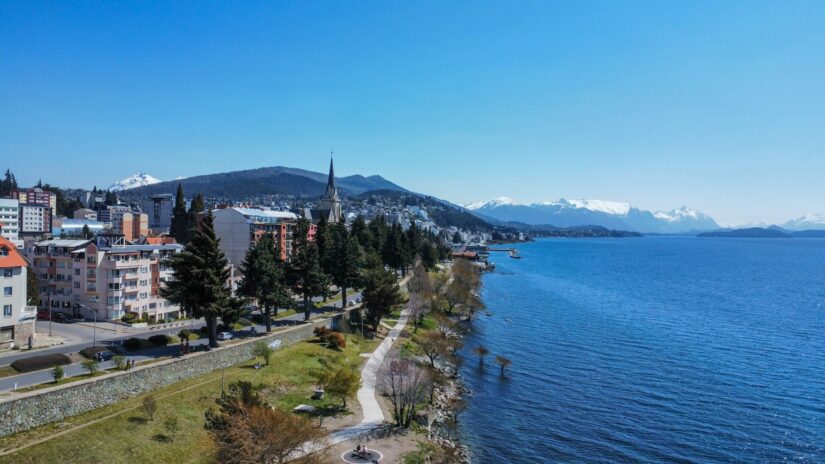 Quantos dias é o ideal em Bariloche? Uma paisagem urbana à beira do lago em Bariloche com edifícios, um caminho arborizado e montanhas - perfeito para onde ficar em Bariloche.