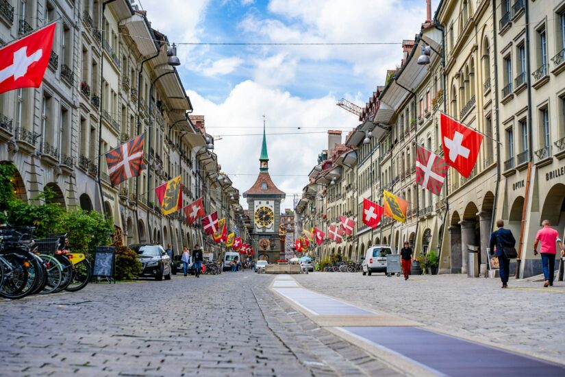 melhores hotéis no Centro Histórico na Suíça Rua de paralelepípedos em Berna, Suíça, com bandeiras, a torre do relógio Zytglogge e dicas sobre onde ficar em Berna nas proximidades.