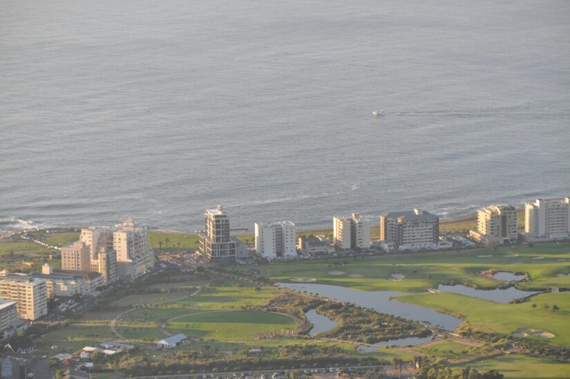 onde ficar em Cape Town no bairro de Green Point na África do Sul Vista aérea de da Cidade do Cabo, edifícios costeiros, campo de golfe verde e o oceano com um pequeno barco à distância.