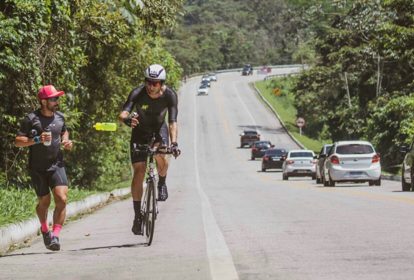 onde ficar em Paraty na Rodovia Paraty-Cunha Um corredor entrega uma garrafa de água a um ciclista na Rodovia Paraty-Cunha ladeada por árvores, com carros passando atrás na Estrada Real no Rio de Janeiro