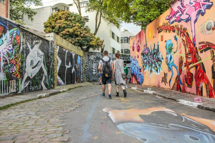 hospedagens na Vila Madalena Duas pessoas caminham pelo Beco do Batman na Vila Madalena repleto de grafites coloridos, explorando onde ficar em São Paulo.