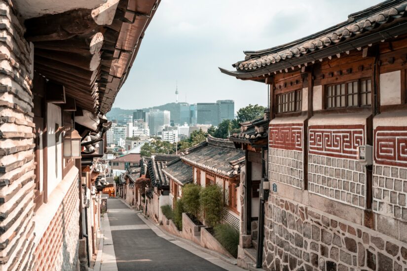 onde ficar em Seul em Bukchon Hanok Village na Coreia do Sul Casas coreanas tradicionais se alinham em uma rua estreita com o horizonte e a torre de uma cidade moderna ao fundo.