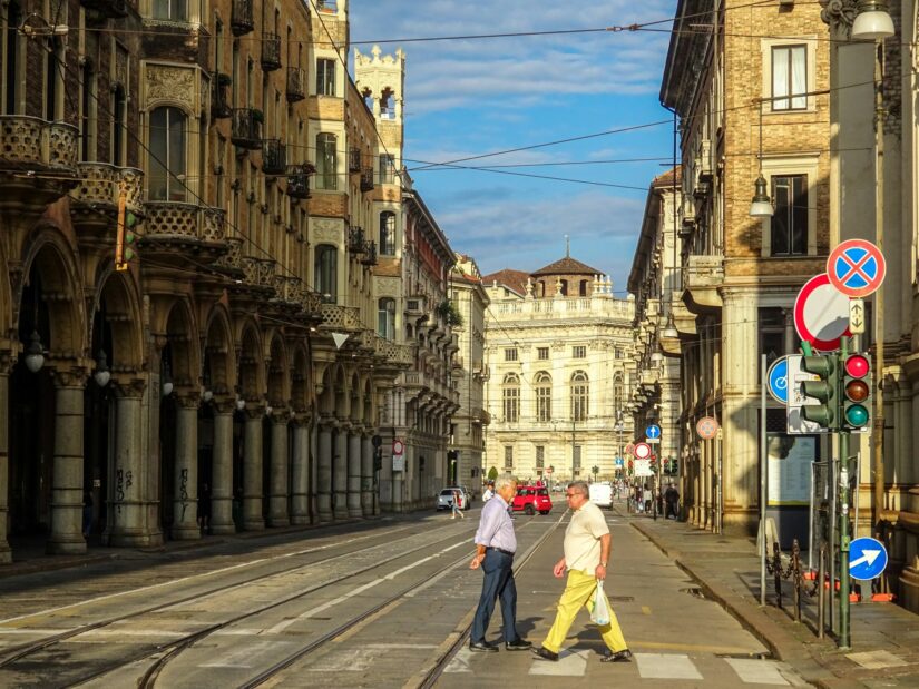 hotéis em Vanchiglia em Torino na Itália Dois homens atravessam uma rua iluminada pelo sol com prédios históricos e trilhos de bonde - perfeito para explorar onde ficar em Turim.