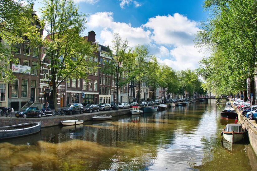 Um canal em Amsterdã com barcos, árvores e onde ficar em Amsterdã entre edifícios tradicionais sob um céu azul.
