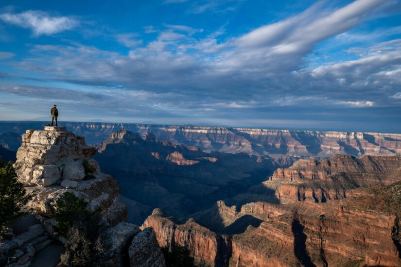 onde ficar no Grand Canyon no North Rim Uma pessoa está em um penhasco rochoso refletindo sobre o North Rim no Arizona, com vista para sua vasta e colorida extensão sob um céu azul.