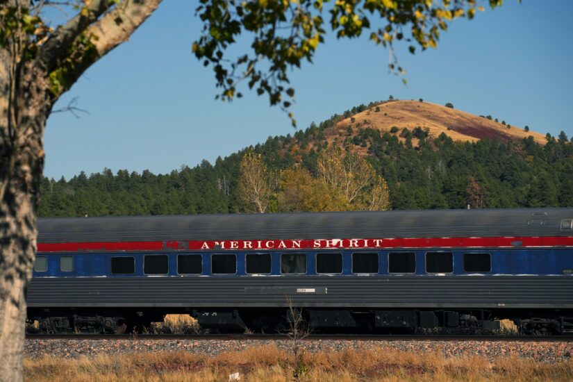 melhores hotéis em Williams no Arizona Um vagão de trem chamado AMERICAN SPIRIT do Grand Canyon Railway passa em Williams perto de onde ficar no Grand Canyon, com colinas, árvores e um céu azul.