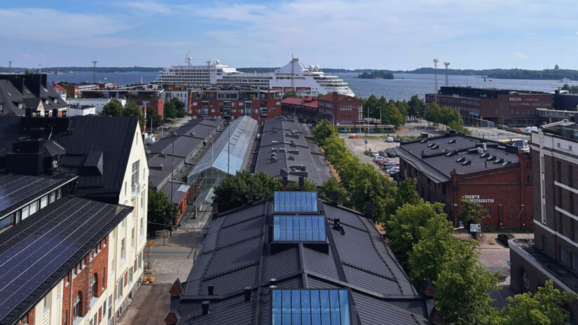 melhores hotéis em Katajanokka na Finlândia Uma paisagem urbana de onde ficar em Helsinque com edifícios de telhado preto, árvores e navios de cruzeiro à beira-mar sob um céu azul no bairro de Katajanokka na Finlândia.