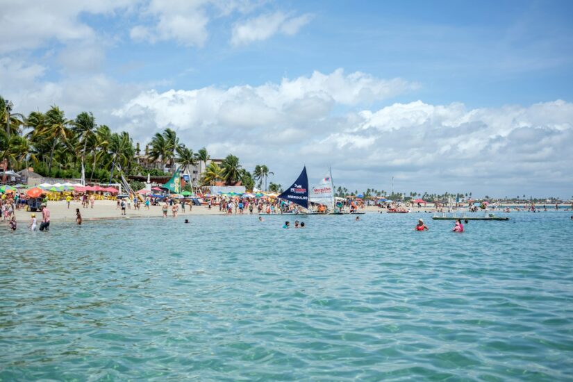 melhores hotéis e pousadas em Ipojuca Praia tropical lotada em Porto de Galinhas com guarda-sóis, palmeiras e barcos em águas azuis claras sob um céu parcialmente nublado. A vila central é uma das melhores regiões onde ficar em Porto de Galinhas.