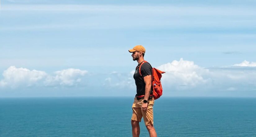 Homem de bermuda e boné com mochila laranja à beira-mar sob um céu azul com nuvens.