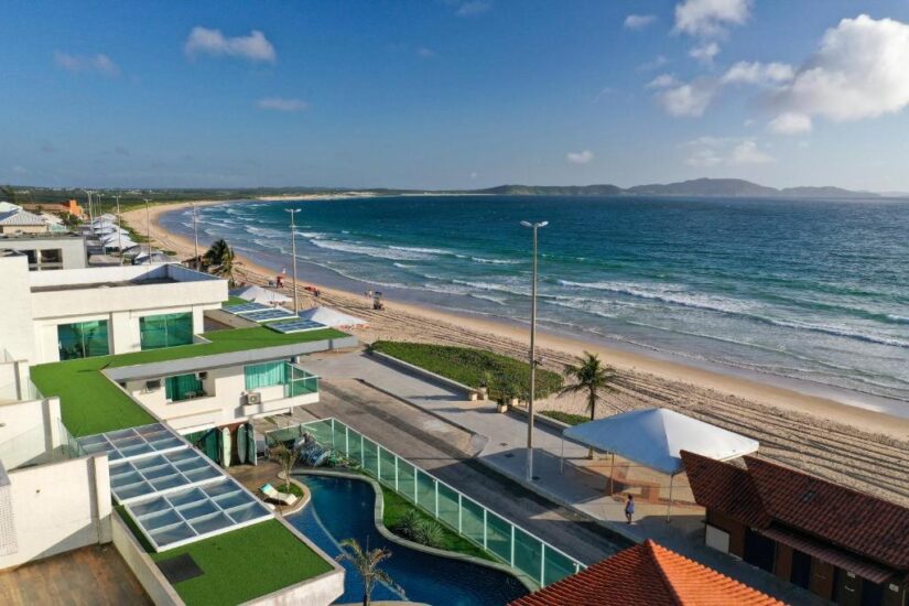 onde ficar em Cabo Frio na Praia do Peró Vista aérea da Praia do Peró no Rio de Janeiro: rua à beira-mar, edifícios modernos, piscina e ondas sob um céu parcialmente nublado.