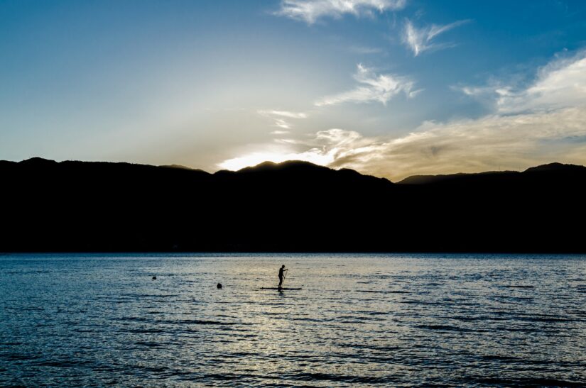 hotéis e pousadas na Praia de Itaquanduba em São Sebastião Pessoa praticando paddleboarding na Praia de Itaquanduba ao pôr do sol, sonhando com onde ficar em Ilhabela, com as montanhas silhuetadas atrás.