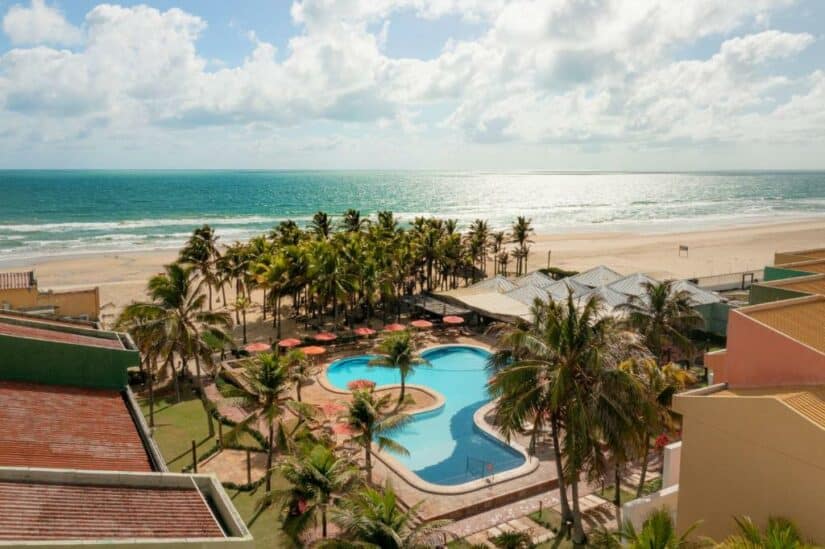 Vista aérea de um resort à beira-mar com piscina, palmeiras e o oceano ao fundo mostra como e onde ficar em Aquiraz.