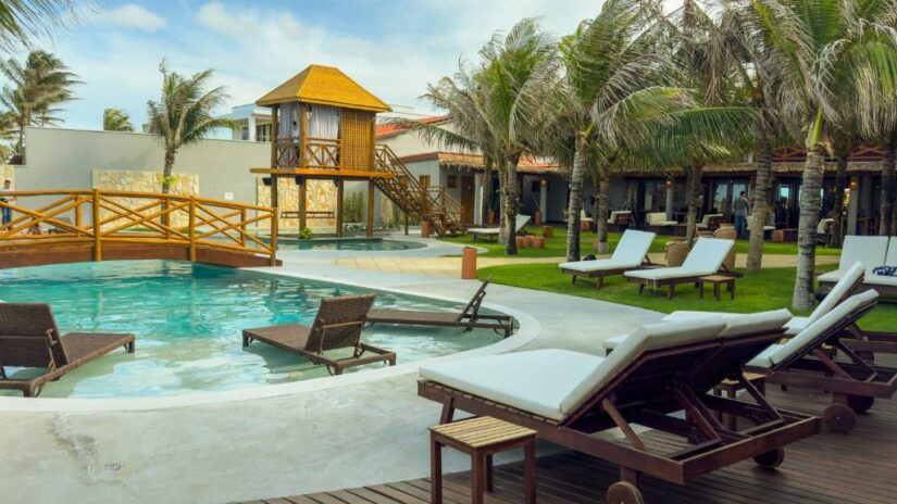 Espreguiçadeiras e palmeiras cercam uma piscina com uma ponte de madeira e uma cabana em um ambiente de resort tropical em Aquiraz.