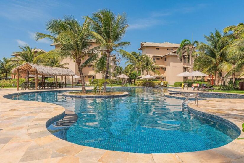 onde ficar em Aquiraz na Praia do Japão no Ceará Piscina externa curva com palmeiras, espreguiçadeiras e guarda-sóis em um resort no Ceará, perto dos prédios bronzeados do resort.