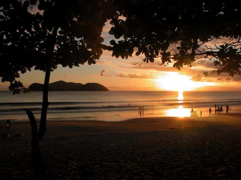 Pôr do sol em uma praia com árvores em silhueta, pessoas na areia e uma ilha à distância mostrando onde ficar em Barra do Una.