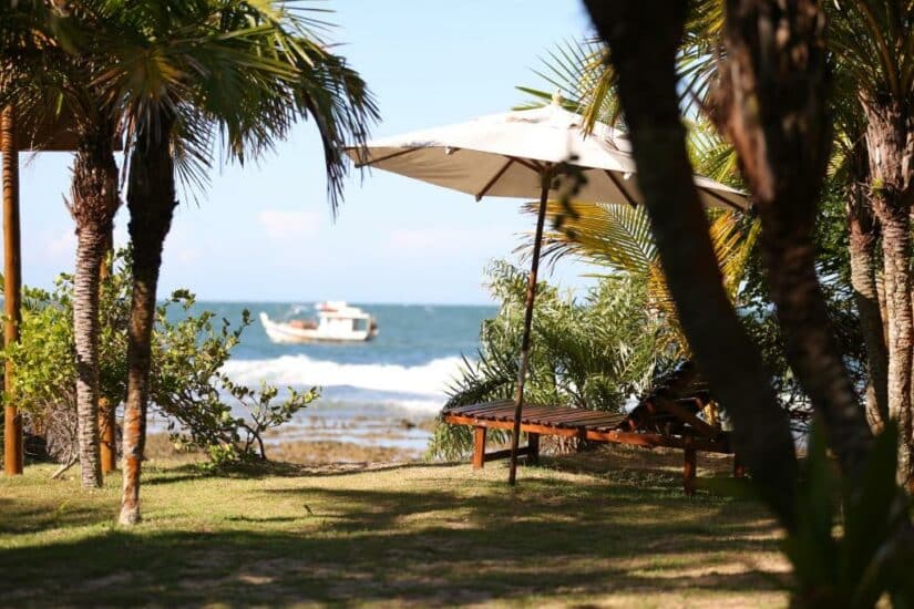 Uma espreguiçadeira com sombra sob um guarda-sol fica de frente para a praia em Maraú Bahia, com um barco no oceano ao fundo.