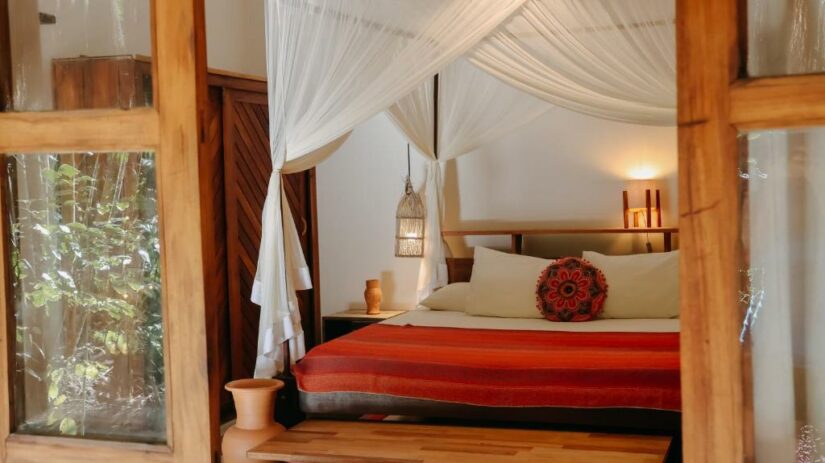 Cama de dossel com cortinas brancas onde ficar em Barra Grande Bahia, cobertor vermelho e laranja e travesseiros decorativos em um quarto aconchegante.