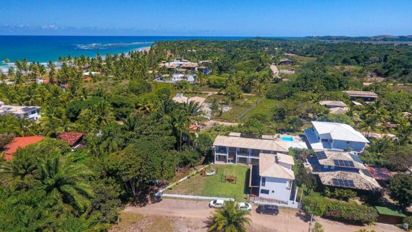 Vista aérea de casas costeiras onde ficar em Barra Grande Bahia cercadas por vegetação exuberante perto de uma praia e do oceano azul sob um céu claro.