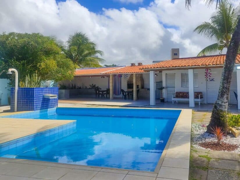 Uma piscina no quintal com azulejos azuis, assentos no pátio, palmeiras e uma casa com telhado vermelho sob um céu nublado mostrando onde ficar em Guarajuba